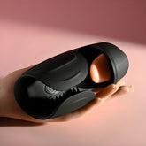 A hand holding the sleek, black Zero Tolerance Lick the Tip Male Masturbator, with its distinctive orange interior design, is set against a soft pink background, highlighting the innovative craftsmanship of this triple-function stroker.